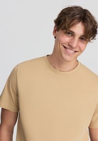 T-shirt beige in cotone a maniche corte con scollatura rotonda. Texture liscia, vestibilità casual. Modello sorridente con capelli castani. Sfondo neutro.