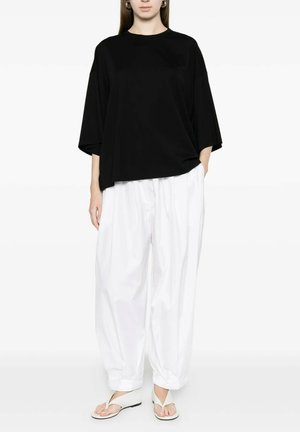 Donna che indossa un ampio top nero largo, pantaloni bianchi larghi e sandali infradito bianchi, in piedi davanti a uno sfondo bianco uniforme.