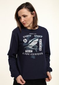 Marineblaue Sweatshirt mit einem Haifischgrafik und dem Text „SNIFF, SNIFF“ in Weiß. Lange Ärmel und Rundhalsausschnitt. Weicher Stoff.
