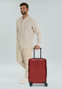 Valise rigide rouge avec un motif en chevrons, roues noires et poignée rétractable, placée à côté d'une personne en vêtements clairs.