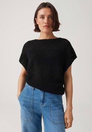 Frau mit kurzem braunem Haar trägt ein schwarzes gehäkeltes Kurzarm-Top und hoch taillierte blaue Jeans, steht mit den Händen in den Taschen.