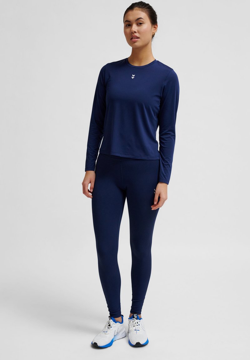 Marineblaues Langarmoberteil mit Rundhalsausschnitt und Logo, kombiniert mit enganliegenden marineblauen Leggings. Model trägt weiße Sportschuhe.