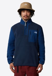 Pullover in pile blu con mezza zip, tasca sul petto e tessuto testurizzato. Abbinato a un cappello a secchiello beige. Sono visibili pantaloni rossi.