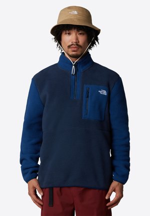 Blå fleece-pullover med kvart glidelås, brystlomme og teksturert stoff. Kombinert med en beige bøttehatt. Røde bukser er synlige.