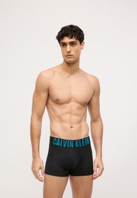 Crne bokserice sa širokim tirkiznim pojasom na kojem je otisnuta oznaka "CALVIN KLEIN". Tkanina izgleda glatko i rastezljivo, čvrsto pristaje.