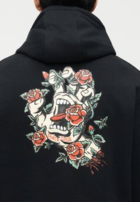 Fekete kapucnis pulóver a hátulján grafikával, amely egy sikoltozó kezet ábrázol, rózsatövisekbe tekerve, piros rózsákkal és a "Santa Cruz" felirattal.