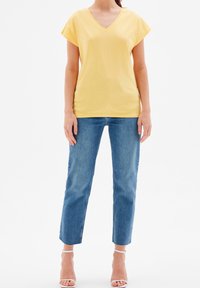 Žluté tričko s výstřihem do V a krátkým rukávem, kombinované s jeansy s rovnými nohavicemi v světlé modré barvě. Oblečení doplňují bílé sandály na pásku.