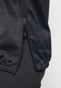 Giacca nera con zip, realizzata in un tessuto liscio e opaco, con orlo elasticizzato e dettagli di cucitura sottili sui polsini. Primo piano dell'area della zip.