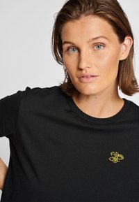 Svart bomullst-shirt med korta ärmar och rund halsringning, med en liten gul broderad bi-design på bröstet.