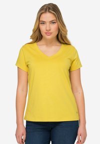 Gele katoenen t-shirt met een V-hals en korte mouwen. Gladde textuur en rechte snit. Draagt met blauwe spijkerbroek.