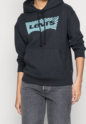 Osoba nosząca czarną bluzę z kapturem Levi's z niebieskim paskowanym logo i szare jeansy, stojąca na jednolitym jasnym tle.