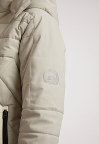Lys beige pufferjakke med diagonalt kviltet mønster, der har en lynlåslomme og et cirkulært emblem på ærmet med bjergdesign.