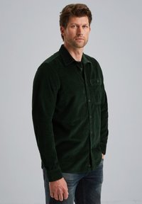 Donkergroene corduroy shirt met een button-down kraag, een borstzak en een gestructureerde stof. Gekleed met donkere jeans, staande tegen een neutrale achtergrond.