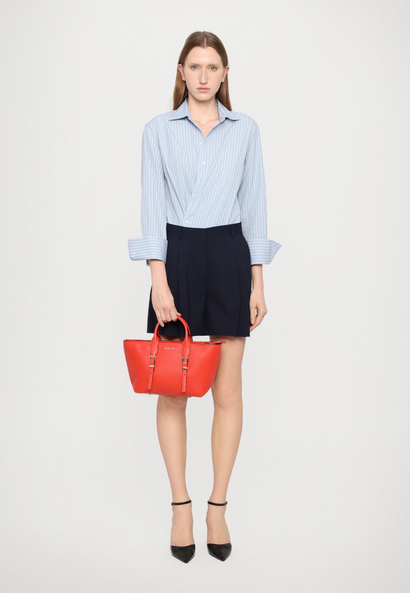Borsa a mano in pelle rossa con manici superiori, superficie texturizzata e accessori in oro, abbinata a una camicia a righe blu e pantaloni corti scuri.