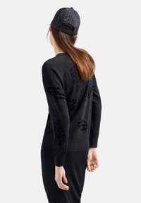 Maglione nero a maglia con motivi neri in rilievo. È indossato un berretto nero abbinato con un design testurizzato. Lo sfondo è bianco semplice.