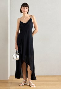 Black asymmetrical dress with cowl neckline, transparent overlay, and silver chain accents on straps. Paired with white sandals and silver bag.