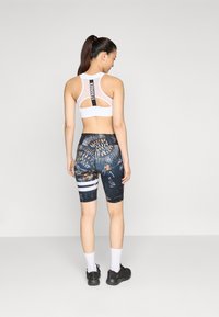 Witte sportbra met mesh achterkant, zwarte en gepatroon gelegde fietsbroek met meerkleurige graphics en witte streep, gedragen met zwarte schoenen en sokken.