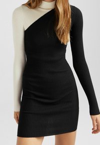 Robe côtelée à col montant, présentant un design en blocs de couleurs : un bras et un côté noirs, un bras et un côté crème, silhouette près du corps.