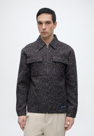Homme portant une veste zippée noir et blanc tachetée avec de grandes poches poitrine et un pantalon beige, debout devant un fond clair uni.