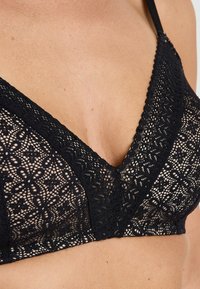 Bralette in pizzo nero con design a V, motivo floreale e bordi scallop. Dotato di spalline regolabili e texture trasparente.