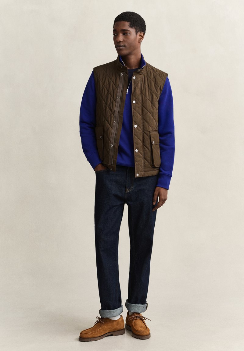 Young man wearing a brown quilted vest over a blue sweatshirt, dark rolled-up jeans, and brown lace-up shoes standing against a plain background.