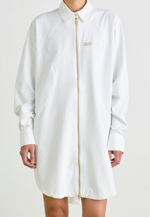 Personne portant une longue veste blanche zippée avec col, fermeture éclair dorée et petit logo sur la poitrine gauche sur un fond uni.