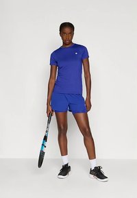 Αθλητής σε μπλε αθλητική ενδυμασία κρατάει μία ρακέτα τένις, φοράει μαύρα αθλητικά παπούτσια και λευκές κάλτσες, μπροστά από έναν απλό φωτεινό φόντο.