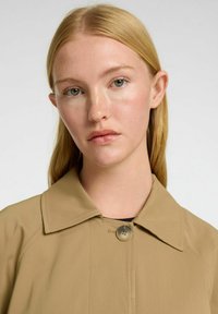 Lys tan jakke med spids krave, der har en frontlukning med knapper, glat tekstur og et minimalistisk design.