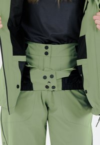 Vihreä hiihtotakki mustilla yksityiskohdilla esillä avoimena, näyttäen istuvan sisäpuolen, napit ja teksturoitua kangasta.