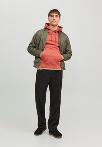 Olijfgroene bomberjack over een koraal hoodie, beige shirt en zwarte broek. Schoeisel is zwart met een bruine zool. Casual gelaagde look.