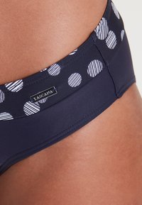 Marinefarbene Schwimmhose mit einem Pünktchenmuster, das durch einheitliche und gestreifte Punkte gekennzeichnet ist. Enthält ein Lascana-Label. Glatte Stofftextur.