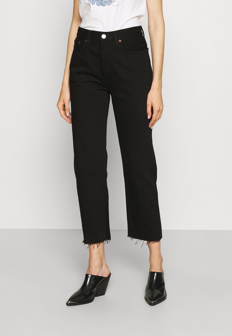Femme portant un jean noir taille haute, droit et court avec des ourlets bruts, un t-shirt blanc à motif graphique et des mules noires à talons pointues.