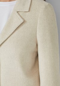 Blazer en mélange de laine beige avec des lapels crantés, une coupe ajustée et une texture douce, mettant en avant un design épuré et minimaliste.