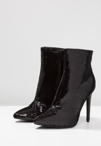 Černé kotníkové boty s flitry, špičatými špičkami a jehlovými podpatky. Mají boční zip pro zapínání a strukturovaný, třpytivý povrch.