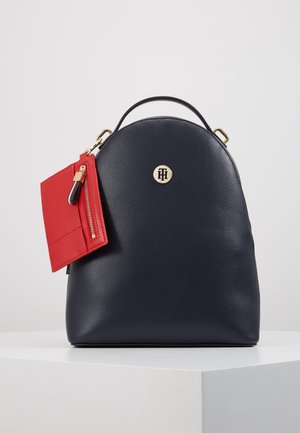 Kompakter dunkelblauer Leder-Rucksack mit Tragegriff oben, goldfarbenem Logo in der Mitte und angebrachtem roten Reißverschlusstäschchen, das an der linken Seite hängt.