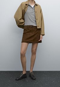 Giacca beige accorciata su un henley grigio, abbinata a una gonna in suede marrone e scarpe con lacci nere. L'outfit presenta una vestibilità comoda e un design semplice.