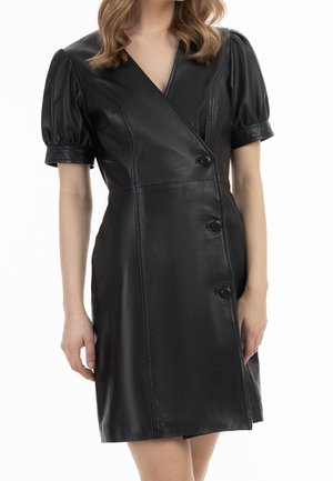 Mujer con un vestido cruzado de cuero sintético negro, con mangas cortas abullonadas y tres botones en la parte delantera.