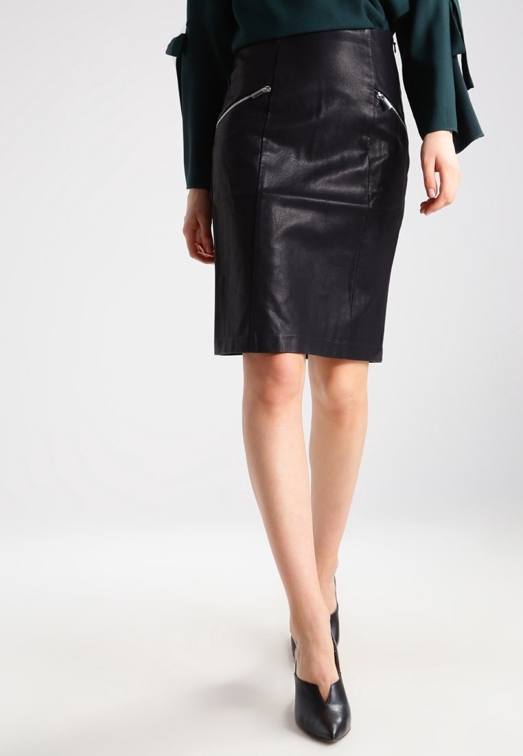 Femme portant une jupe crayon en cuir noir avec des zips à l'avant, associée à des escarpins noirs à bout pointu et un haut à manches longues vert foncé.