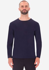 Uomo con barba che indossa una semplice camicia a maniche lunghe di un blu navy scuro, in piedi contro uno sfondo bianco, rivolto in avanti.