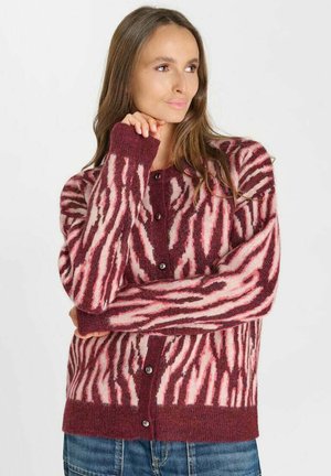 Vrouw met lang bruin haar, die een roze en bordeaux zebraprint gebreid vest met knopen en een blauwe spijkerbroek draagt, kijkt opzij met haar hand bij haar gezicht.