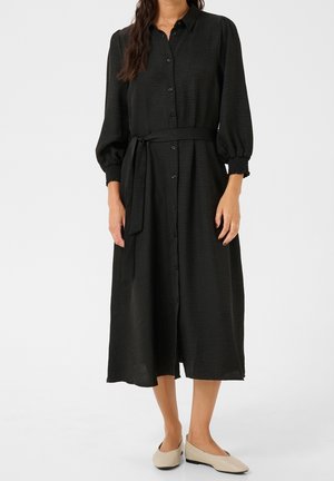 Femme portant une robe midi noire boutonnée avec des manches 3/4 bouffantes, une ceinture nouée à la taille et des chaussures plates beiges, debout devant un fond blanc.