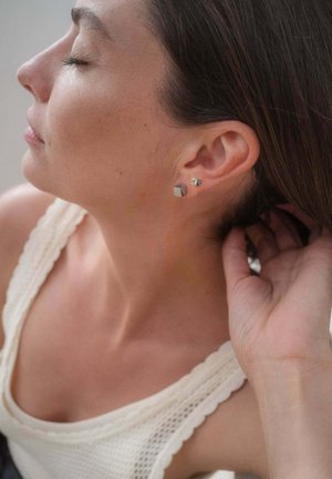 Frau mit geschlossenen Augen, trägt zwei silberne geometrische Ohrstecker, berührt ihr Haar hinter dem Ohr, gekleidet in einem cremefarbenen Stricktop.