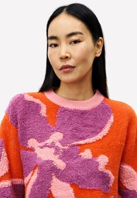 Vrouw met lang zwart haar die een oranje en paarse gestructureerde trui met een bloemenpatroon draagt, recht vooruit kijkend met een neutrale uitdrukking.