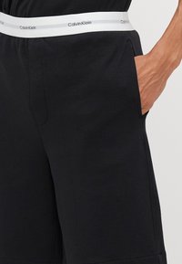 Crne kratke hlače od mekog materijala s bočnim džepovima i bijelim elastičnim pojasom s ponavljajućim logotipom Calvin Klein.