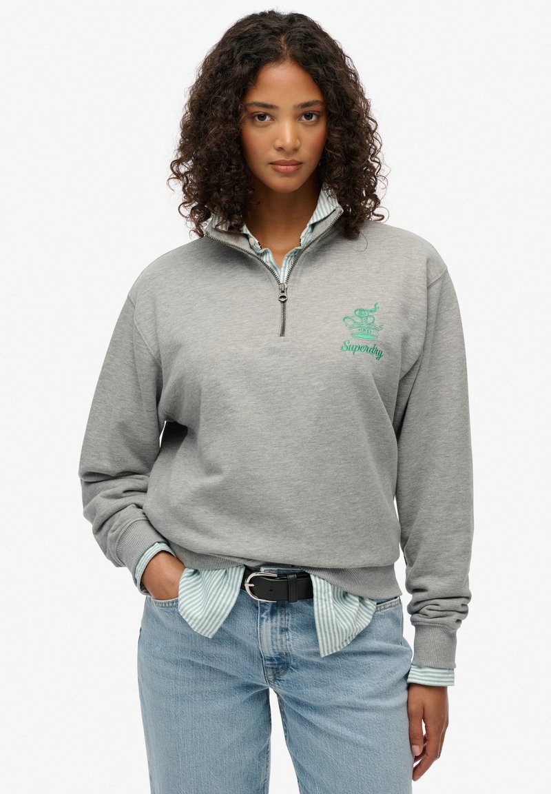 Sudadera gris de forro polar con media cremallera y logo bordado en verde, llevada sobre una camisa a rayas azul. Presenta un corte relajado y puños acanalados.