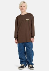 Brązowa koszulka z długim rękawem z małą grafiką na piersi, zestawiona z luźnymi niebieskimi dżinsami i czarnymi trampkami z białymi podeszwami.