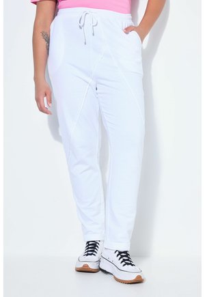 Osoba nosící bílé tepláky se zavazováním, bílé tenisky s černými tkaničkami a růžový top, stojící na bílém pozadí.