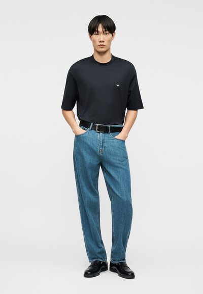Άνδρας που φοράει μαύρο κοντομάνικο μπλουζάκι με λογότυπο, μπλε ψηλόμεσο τζιν, μαύρη ζώνη και μαύρα παπούτσια, στέκεται με τα χέρια στις τσέπες.