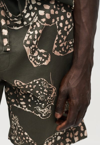 Χέρι με σκουρόχρωμο δέρμα που ακουμπά κοντά σε ελιάς πράσινα σορτς με εκτύπωση μεγάλων ανοιχτόροζ προσώπων λεοπάρδαλης και πουά μοτίβα.