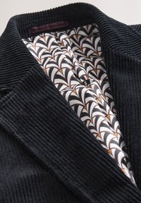 Schwarze Cordjacke mit breiten Revers, die ein geometrisch gemustertes Seidenfutter in Weiß, Schwarz und Braun im Krageninnenbereich zeigt.